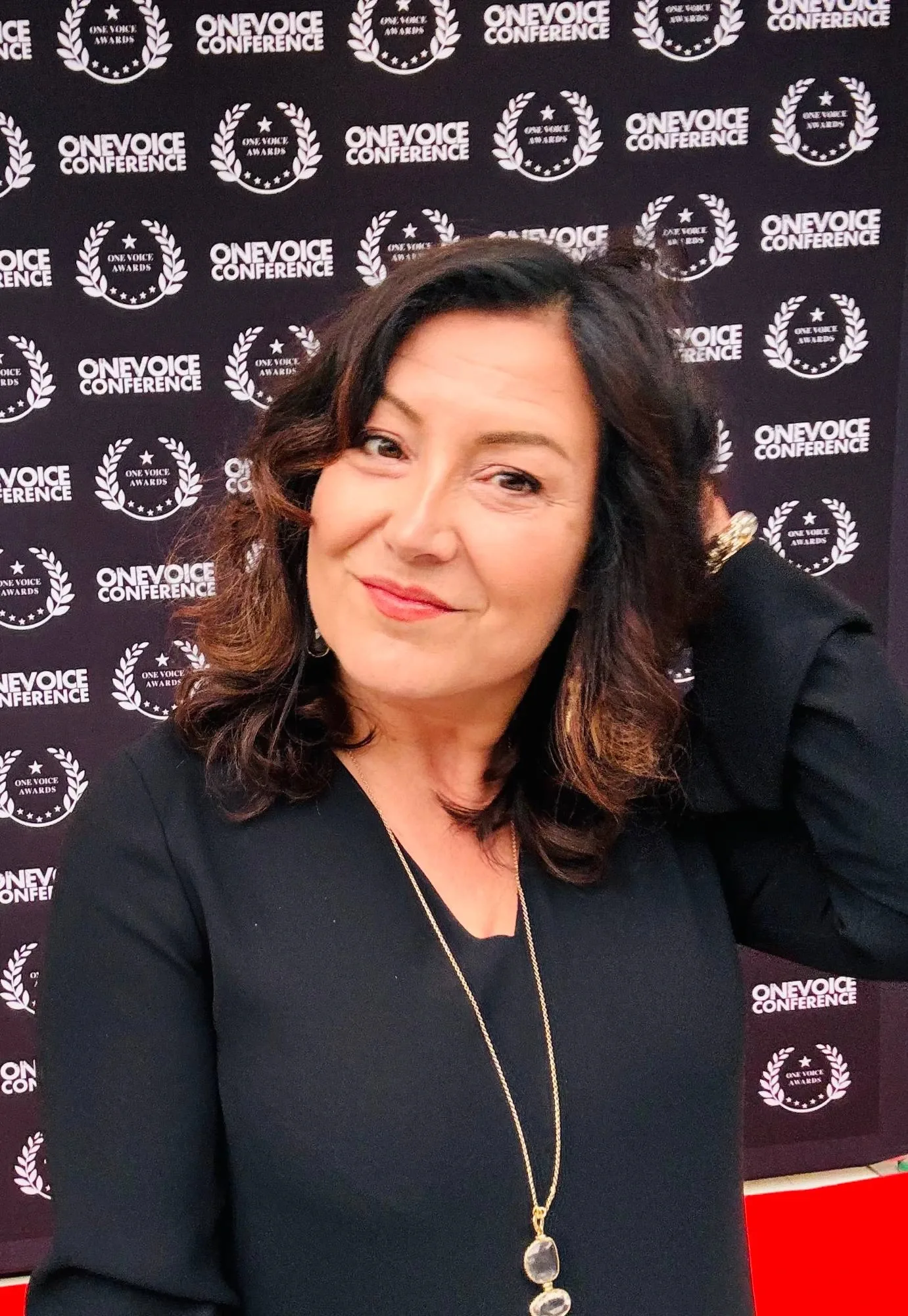 Estelle Hubert en tenue noire, une main dans les cheveux, souriante devant le mur de presse de la One Voice Conference